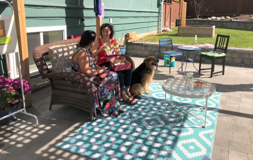 Shokhan and Alysha sit on patio furniture in dappled light below a patio arbour, accompanied by a large dog.