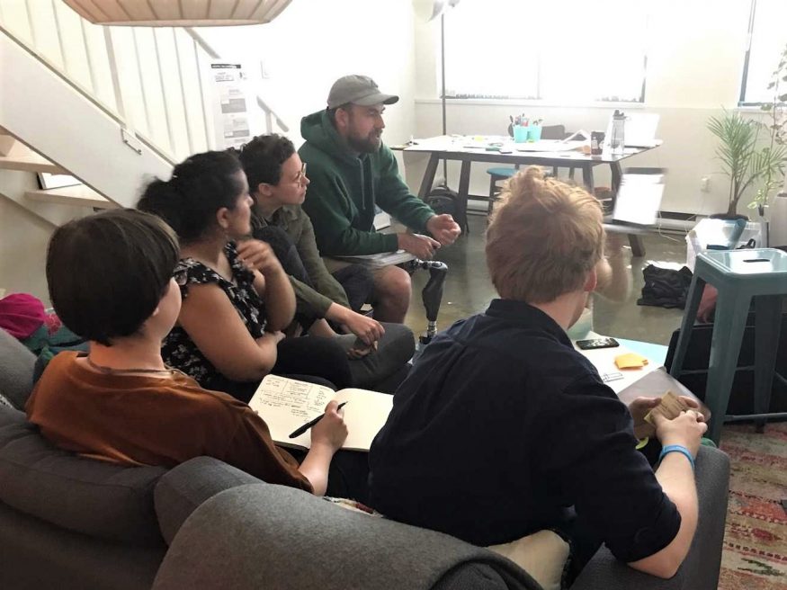 The team seated on a couch all looking to the front of the room, getting a lesson.