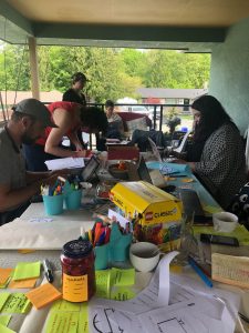 Team members sit on both sides of a long table covered in materials and tools, head down, deeply focused.