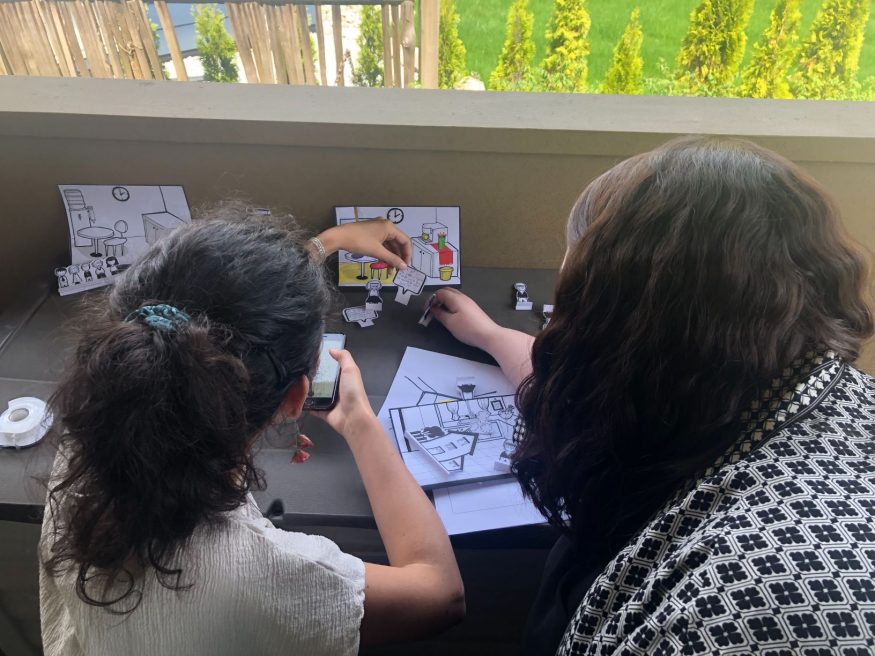 Photo taken from over the shoulders of two women who are moving around small paper props in a tiny box scene.