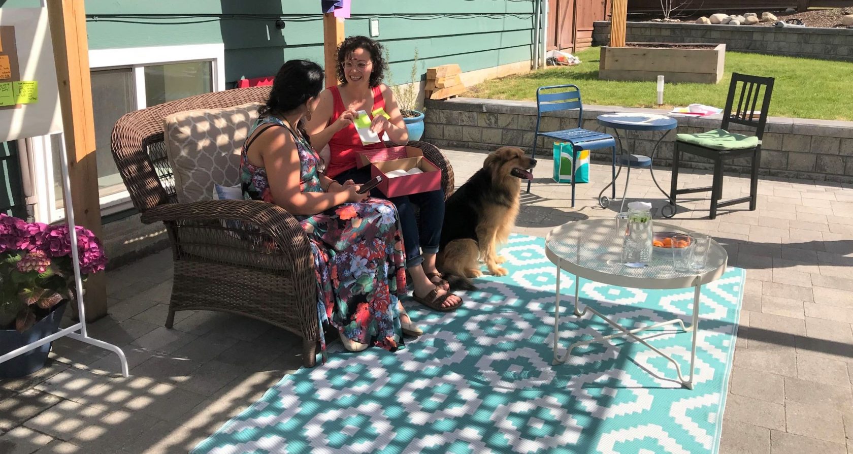 Shokhan and Alysha sit on patio furniture in dappled light below a patio arbour, accompanied by a large dog.