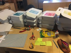 Boxes piles high on a desk strewn with glue, scissors, ruler, self-healing mat, tape, and felts.