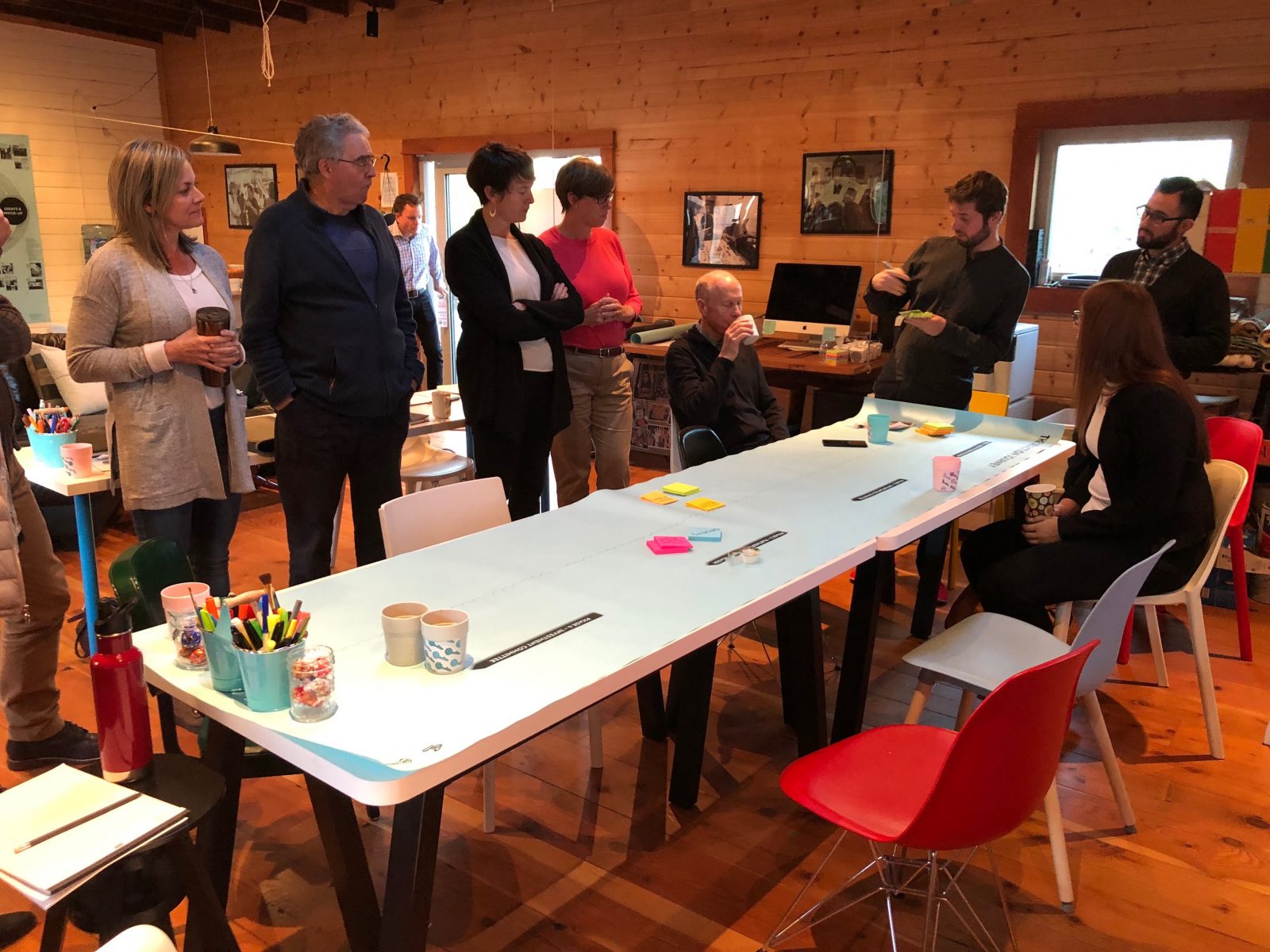 Eight adults sit and stand around a long table laid out with paper, post-its and pens.