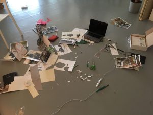 A laptop, cutting mat, several exacto knives, cut-out paper, jar of pens, and scraps of paper, spread messily around a concrete floor.