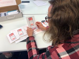 Photo taken from over the shoulder of a long-haired, bespectacled young man, looking through box content, deciding what kind of person he would choose to occupy one of the roles.