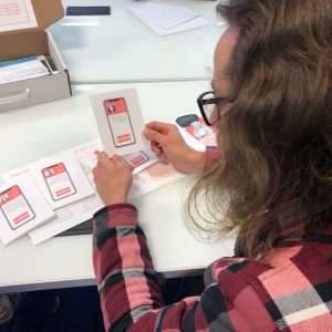 Photo taken from over the shoulder of a long-haired, bespectacled young man, looking through box content, deciding what kind of person he would choose to occupy one of the roles.