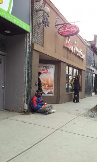 A Man in a red and black jacket and toque sits on the sidewalk in front of Tim Hortons, holding a paper cup.