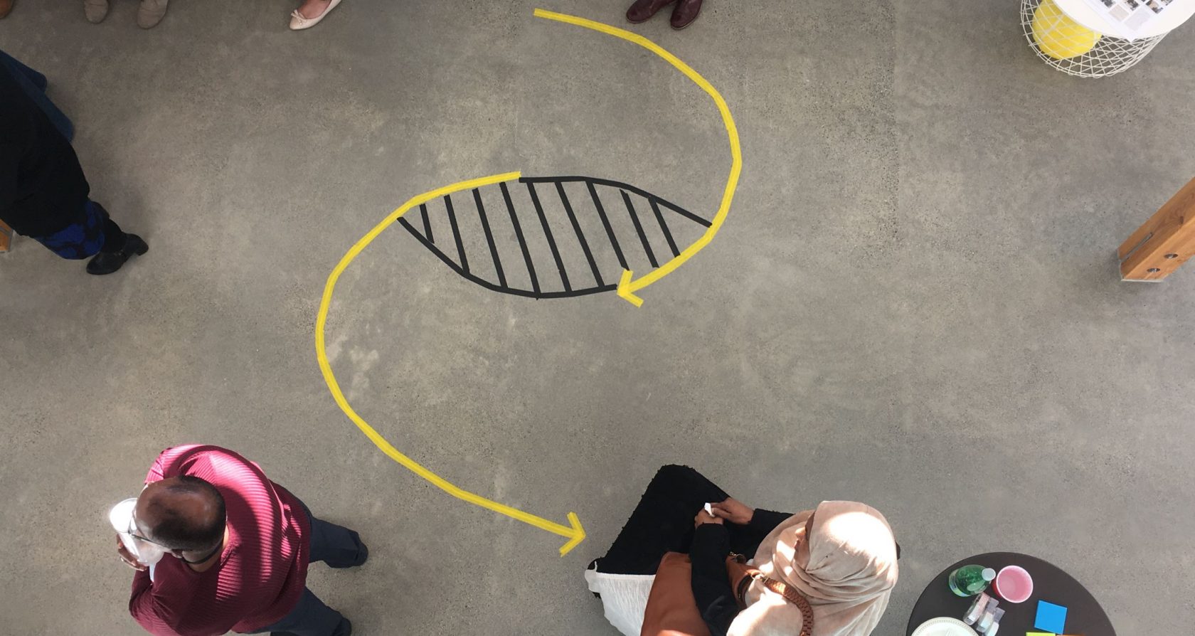 A concrete floor with a taped diagram of living and dying systems (two arced arrows in an 's' like formation but with a gap in the middle of the 's' that looks like an eye. People mill about.