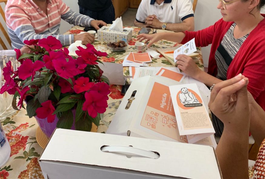 Four people sit round a table (heads not shown) looking through materials in boxes"