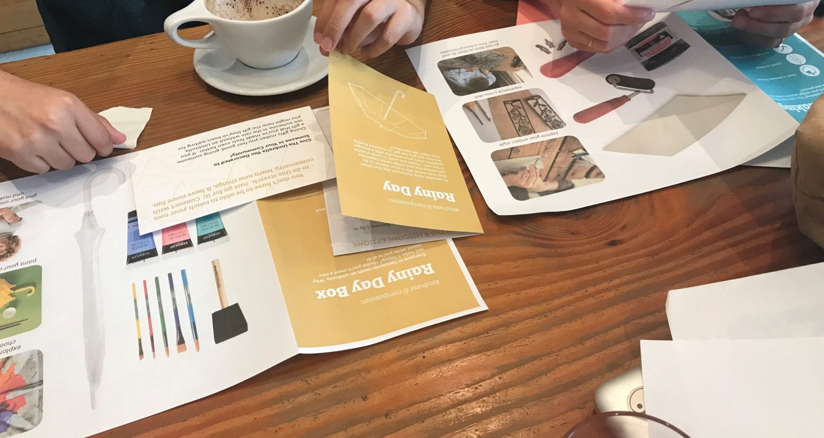 Materials illustrating the Rainy Day Box activity are spread on a table in front of two people whose hands are visible.