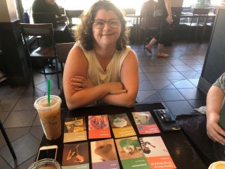 An end user smiles proudly with arms crossed over her chest, leaning on a cafe table that contains a grid of colourful cards with various titles and a frozen coffee drink.