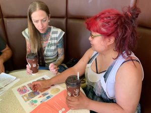 Two women sitting in a booth, one with long, straight blond hair, tattoos on her exposed neck, collar bone, and upper arms. The other has dyed bright red hair, and is wearing overalls. Both are holding a paper McDonald's coffee cup in their hands, resting on the table. They are looking down at colourful paper materials.