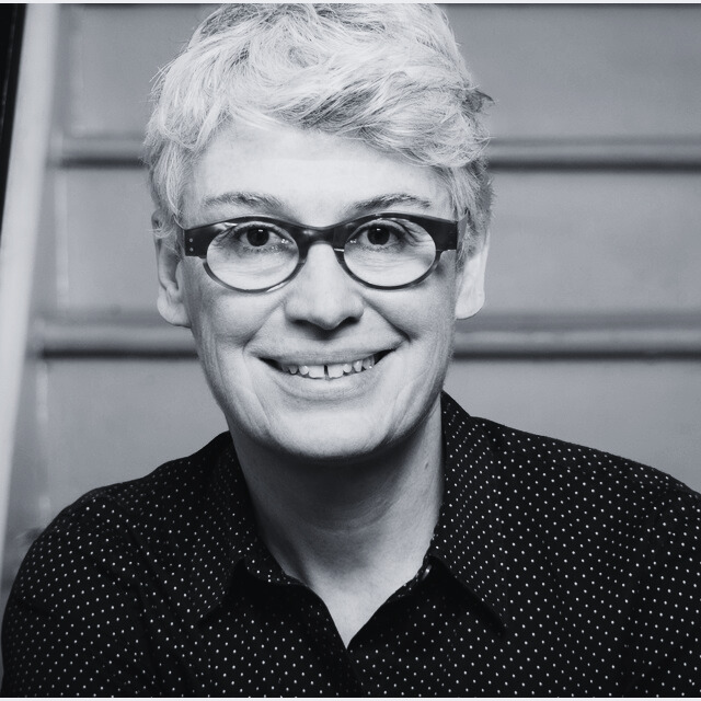A black and white portrait image of a woman with short grey hair, and horn-rimmed glasses, wearing a button-up shirt, and smiling, seated on stairs.