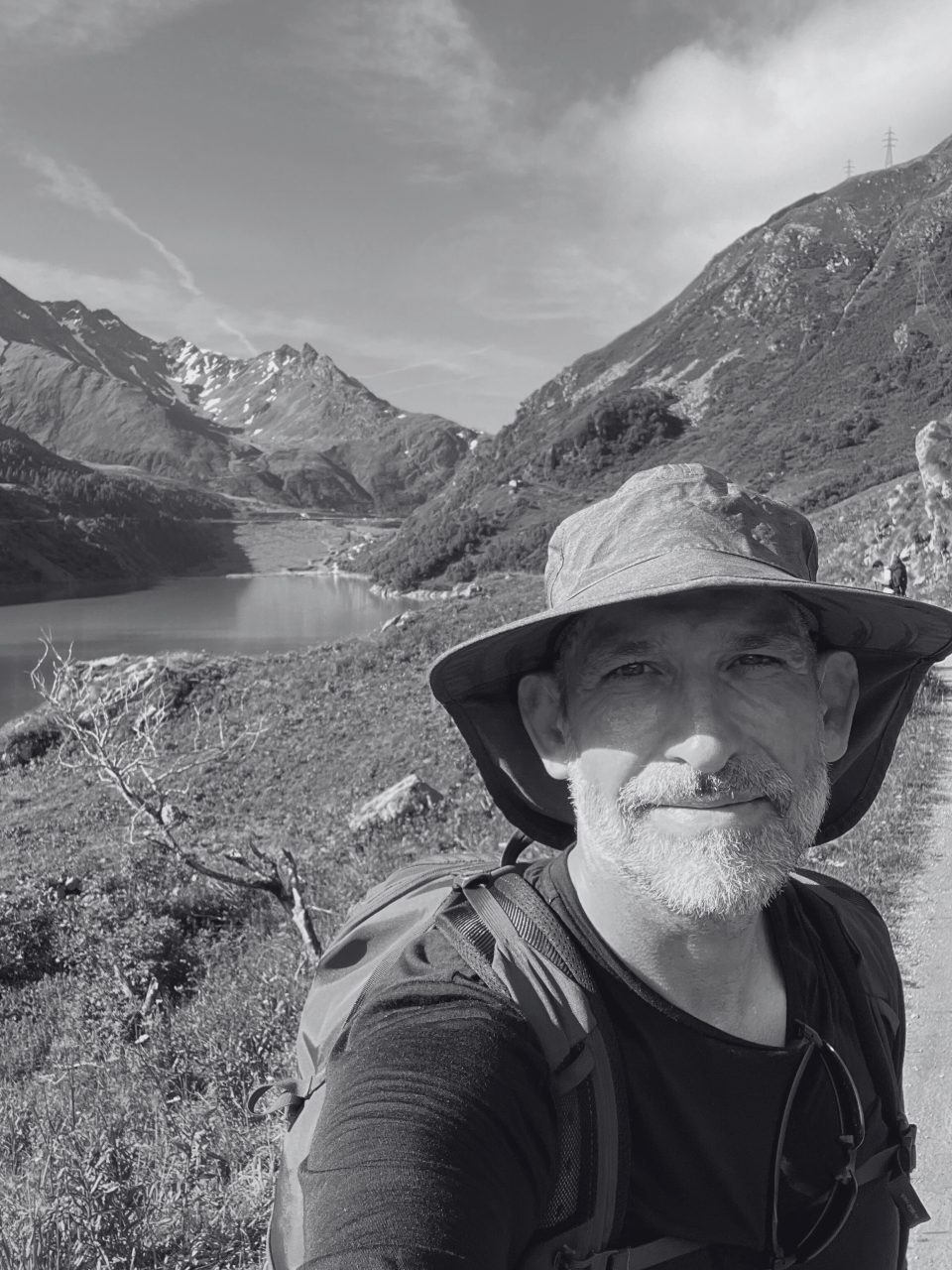 Gord's bearded face is shaded by a wide-brimmed hat. He wears a red backpack over a black tshirt, and looks up at the camera against a panoramic backdrop in the Alps