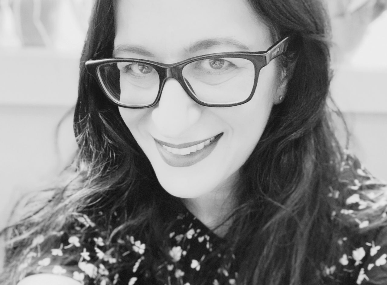 A close-up of a smiling White woman in black-rimmed glasses with long, dark hair fraiming her face, looking up towards the camera.